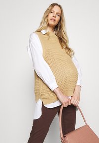 Um colete bege de malha sobre uma camisa branca, combinado com calças castanhas e uma mala texturizada rosa. O colete tem um design solto e de trama aberta.