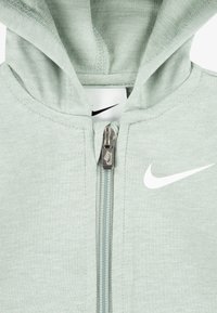 Šviesiai žalia Nike zip-up gobtuvinė striukė su sidabriniu užtrauktuku, balta logo krūtinėje ir tekstūruota medžiaga, su gobtuvu ir priekiu užtrauktuku.