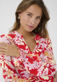 Piros virágmintás blúz, rózsaszín és sárga részletekkel, V-nyakkal és redőzött ujjal; könnyű, selymes anyag, puha tapintással.
