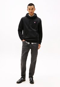Schwarzer Kapuzenpullover mit einer Fronttasche, kombiniert mit grauen Jeans und einem karierten Hemd darunter. Das Model trägt schwarze Schuhe.