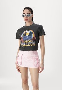 Tricou grafic negru cu imprimeu colorat și text, asociat cu o fustă mini din denim roz cu buzunare tip cargo și fermoare. Ochelari de soare albi.