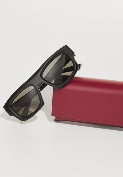 Occhiali da sole neri con un design geometrico, lenti scure e dettagli dorati, accanto a un astuccio in pelle bordeaux impresso con "Gucci".