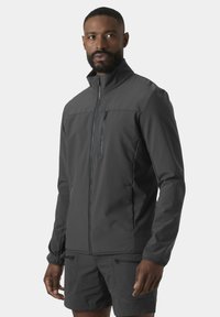 Hombre con chaqueta gris oscuro con cierre y pantalones cortos a juego de pie contra un fondo liso, mirando ligeramente hacia un lado.