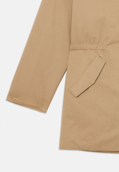 Μπεζ μανίκι παλτού και μπροστινή τσέπη με διαγώνια λεπτομέρεια στην καλύπτρα σε λείο υφασμάτινο υπόβαθρο.