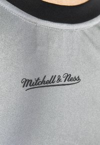 Tissu de maillot de sport gris avec une finition texturée. Présente un col rond noir et un logo brodé "Mitchell & Ness" en noir.