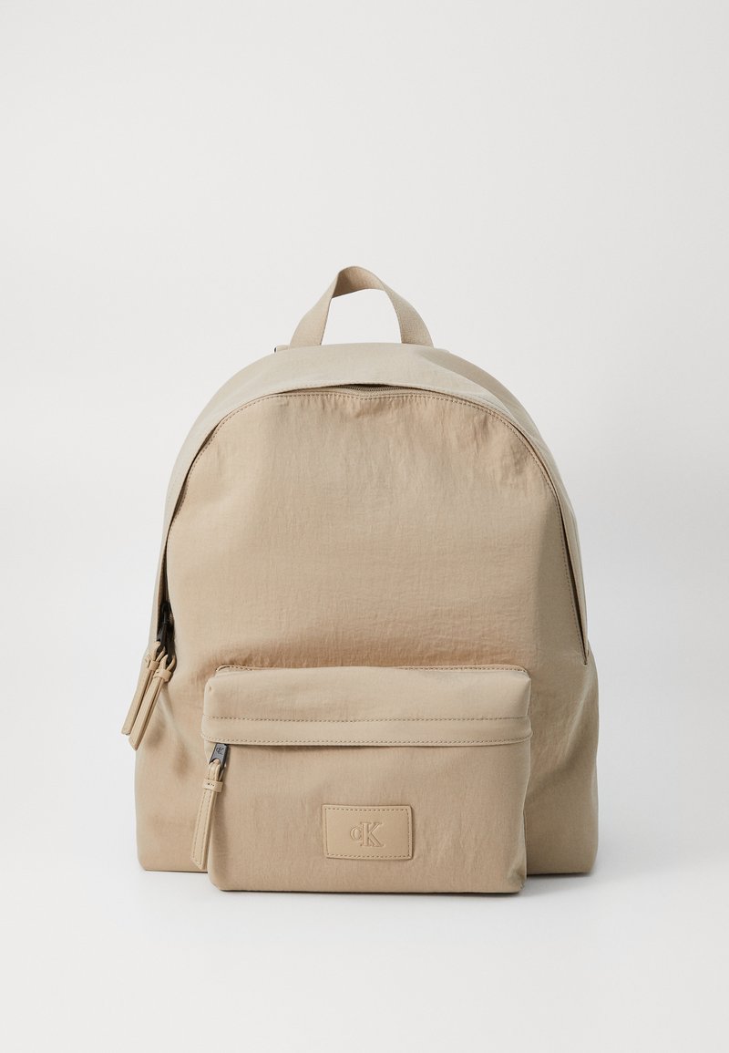 Zaino in nylon beige con forma arrotondata, dotato di una tasca frontale con zip, doppie maniglie superiori e una finitura testurizzata sottile.