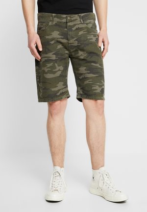 Personne portant un short mi-long à motif camouflage vert, un haut noir et des baskets montantes blanches, debout devant un fond clair uni.