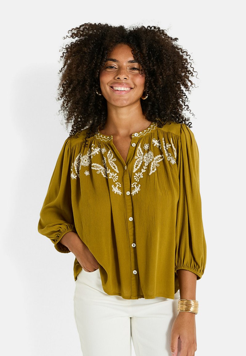 Blouse vert moutarde avec de longues manches ballons, fermeture à boutons sur le devant et broderie florale blanche sur la poitrine. Tissu léger et texturé.