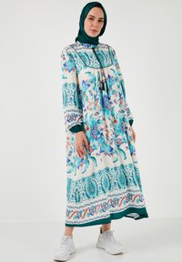 Long, loose-fitting dress in white with intricate blue and turquoise floral patterns. Features long sleeves and a drawstring neckline.