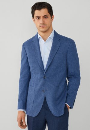 Texturerad blå blazer med hounds-tooth mönster, två framfickor, sned skärning på kragen och enkel knappstängning, kombinerad med en ljusblå skjorta.