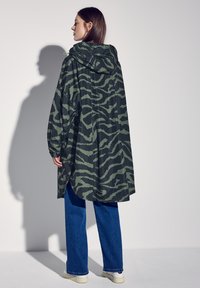 Grön camouflage-mönstrad regnrock med huva, lös passform, lättviktsmaterial, långärmad och med sidosprund, kombinerad med blå vida jeans.