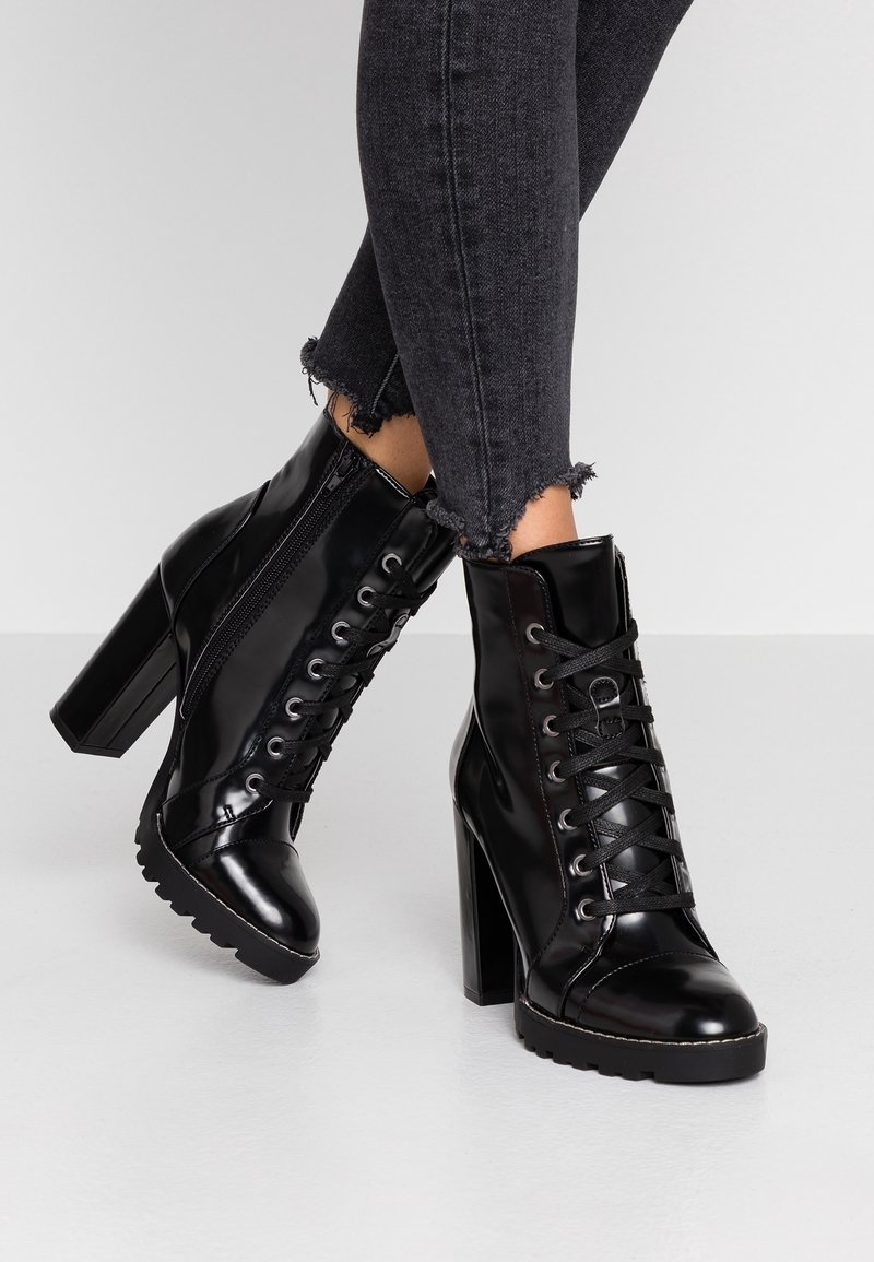 Bottines à talons hauts en cuir verni noir avec lacets et fermeture éclair latérale, portées avec un jean skinny gris foncé à ourlet effiloché.