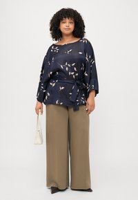 Top in satin blu navy con motivo floreale, maniche ampie e cintura a fiocco, abbinato a pantaloni a gamba larga verde oliva a vita alta e una piccola borsa bianca.