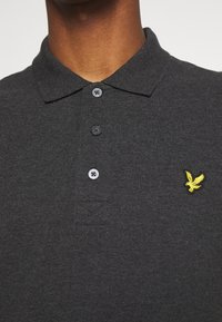 Šedé polo tričko vyrobené z texturované látky s límečkem, s třemi černými knoflíky a žlutou vyšitou logem orla na hrudi.