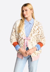 Strukturierte cremefarbene Strickjacke mit braunen Punkten, pinker Krawattenverschluss, orangefarbenen und blauen Rippbündchen, getragen über einem blau-weißen gestreiften Hemd.