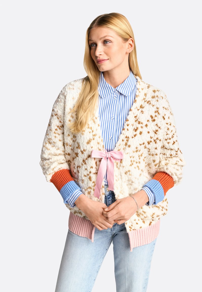 Strukturierte cremefarbene Strickjacke mit braunen Punkten, pinker Krawattenverschluss, orangefarbenen und blauen Rippbündchen, getragen über einem blau-weißen gestreiften Hemd.