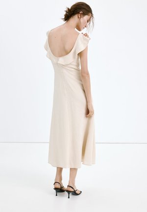 Femme portant une robe midi crème sans manches avec un détail volanté aux épaules et un dos ouvert, associée à des sandales à talons noires à brides.