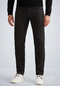 Man in een donkergrijze slim-fit broek en witte sneakers, staand op een lichtgrijze vloer tegen een effen achtergrond.