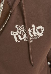 Felpa con cappuccio marrone caratterizzata da un design grafico bianco con lettere stilizzate sul davanti, tessuto texturizzato e chiusura con zip.