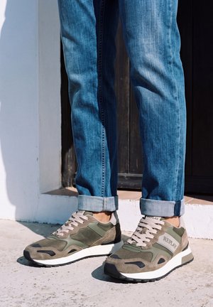 Jean bleu à revers et baskets Replay gris olive à semelles blanches, portés à l’extérieur sous un soleil éclatant.