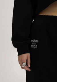 Zwarte trainingsbroek van een gestructureerde stof met een elastische tailleband. Witte tekstdetails op het broekbeen omvatten merk- en bewegingsbeschrijvingen.