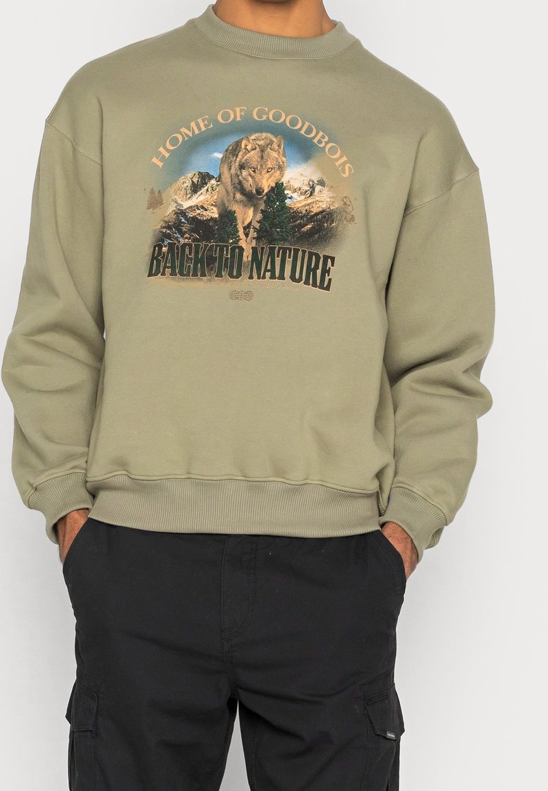 Homme portant un sweat-shirt vert olive avec un loup, des arbres, des montagnes et le texte "Home of Goodbois Back to Nature" sur le devant.