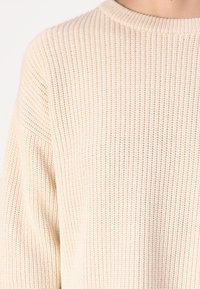 Suéter beige de punto con textura acanalada, cuello redondo y hombros caídos; cuenta con un ajuste holgado y tela suave y cálida.