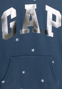 Felpa con cappuccio in tessuto blu scuro, caratterizzata da grandi lettere metalliche argento "GAP" e motivi di stelle bianche sparse; include una tasca frontale.