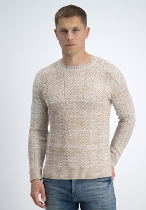 Male model wearing beige textured knit sweater with geometric patterns and blue jeans, standing against plain light background.