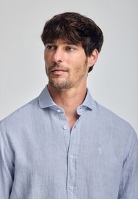 Jasnoniebieska lniana koszula zapinana na guziki z kołnierzykiem, z małym wyszywanym logo na lewej piersi oraz gładkim, teksturowanym materiałem.