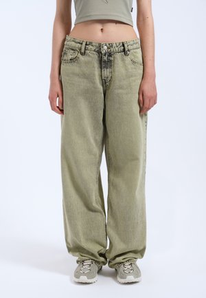 Jean en denim surdimensionné vert clair avec une texture décolorée et une fermeture à bouton à l'avant, associé à des chaussures de sport grises. Pas de motifs visibles.