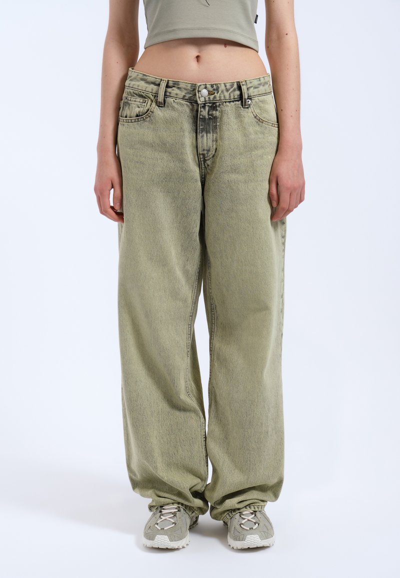 Světle zelené oversized džíny z denimu s jemně vybledlým vzhledem a předním zapínáním na knoflíky, doplněné šedými sportovními botami. Žádné viditelné vzory.