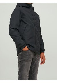 Osoba w czarnej kurtce zapinanej na zamek z elastycznymi mankietami, z jedną ręką w kieszeni, w połączeniu z szarymi dżinsami na jednolitym tle.