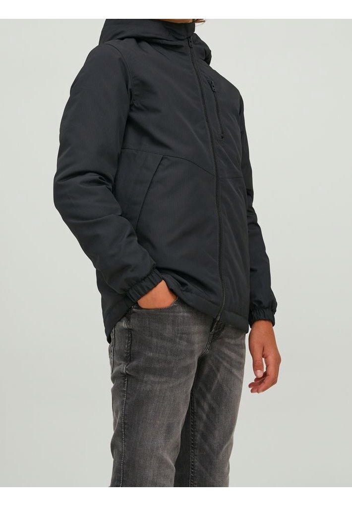 Osoba w czarnej kurtce zapinanej na zamek z elastycznymi mankietami, z jedną ręką w kieszeni, w połączeniu z szarymi dżinsami na jednolitym tle.