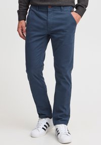 Chino bleu marine pour hommes en mélange de coton, avec une coupe droite, deux poches latérales et une fermeture à bouton. Porté avec des baskets blanches.
