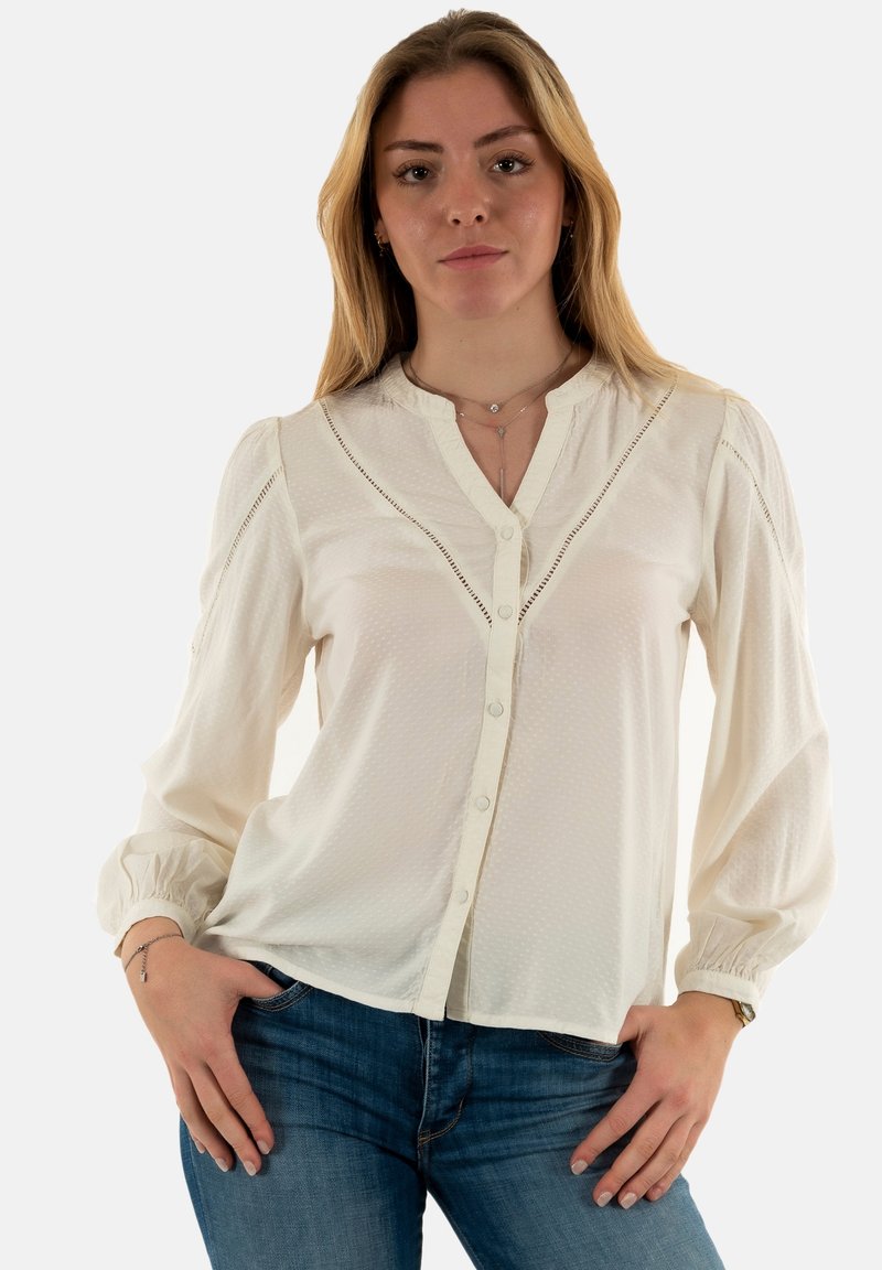 Blusa blanca de botones con un tejido texturizado, escote redondo y mangas abullonadas. Presenta un bordado ornamentado a lo largo de las costuras del cuerpo.