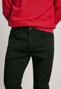 Black denim jeans with a slim fit, featuring five pockets, a front button closure, and subtle stitching details. Paired with a red knit sweater.