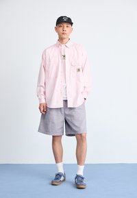 Camisa de botones de color rosa claro con bolsillos, combinada con pantalones cortos de rayas grises texturizadas. Una gorra negra y zapatillas azules completan el conjunto.
