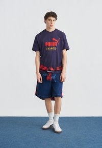 Νέος άνδρας που φοράει μπλε σκούρο T-shirt Puma King, σορτς από τζιν, λευκές κάλτσες, λευκά αθλητικά παπούτσια και κόκκινο πουκάμισο καρό δεμένο γύρω από τη μέση, στέκεται σε μπλε χαλί.