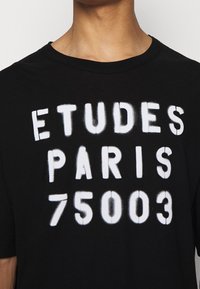 Black cotton t-shirt with white spray-painted text: "ETUDES PARIS 75003." Crew neckline, short sleeves, regular fit.