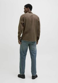 Brun långärmad skjorta med krage och knäppningar vid ärmsluten, kombinerad med ljusblå jeans och svarta skor, sedd bakifrån.