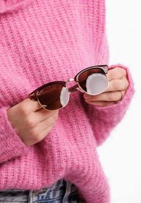 Sunglasses ve tvaru kočičích očí s tmavě hnědým tónováním, zlatými akcenty a průhlednými rámy, drženy proti texturovanému růžovému svetru.