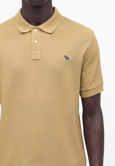 Homme portant un polo beige à manches courtes avec deux boutons et un petit logo zèbre brodé sur le côté gauche de la poitrine.