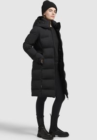 Zwarte lange pufferjas met een capuchon, gewatteerd ontwerp, zijzakken, gedragen over een zwarte top en legging, gecombineerd met zwarte enkellaarsjes en een beanie.
