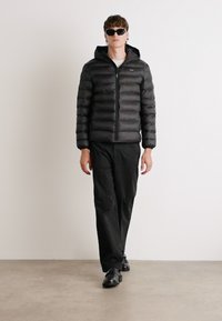 Giacca a vento nera con cappuccio e cuciture orizzontali, abbinata a pantaloni e scarpe nere, indossata da una persona in un ambiente minimalista.
