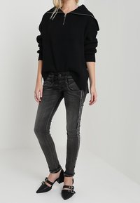 Suéter negro de punto acanalado con cuello de media cremallera, combinado con jeans pitillo gris oscuro con detalles de costuras laterales y tacones negros de punta puntiaguda.