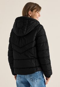 Sort pufferjakke med hætte, der har et chevron-syet mønster, ribbede detaljer og struktureret stof, båret med lyseblå jeans.