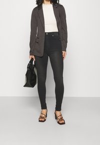 Zwarte high-waisted skinny jeans, een licht geribd topje, een donker colbert en sandalen met hak en bandjes; een zwarte tas met structuur vasthoudend.