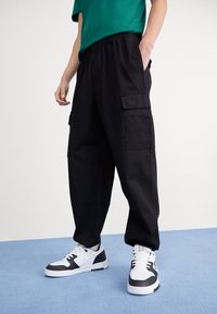 Mustat cargo-housut, joissa on joustava vyötärönauha ja sivutaskut, yhdistettyinä mustavalkoisiin lenkkareihin. Kangas vaikuttaa kestävältä ja sileältä.