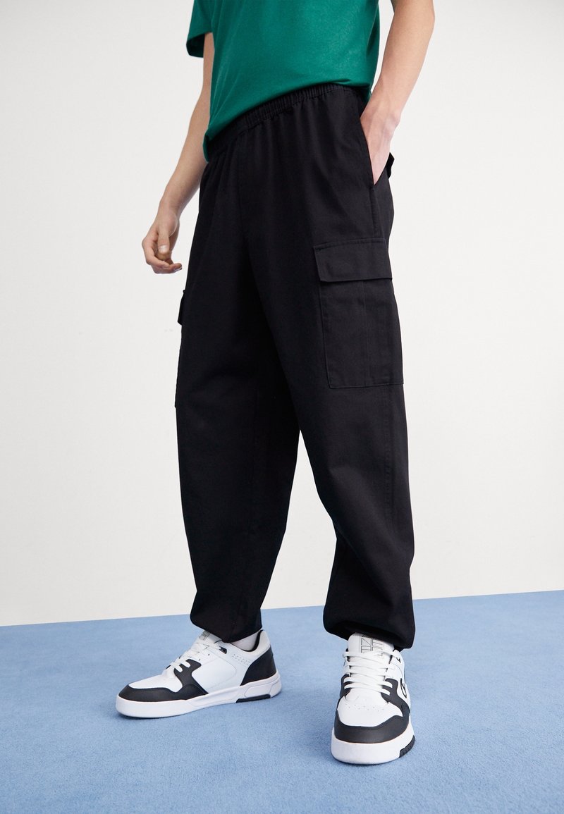 Mustat cargo-housut, joissa on joustava vyötärönauha ja sivutaskut, yhdistettyinä mustavalkoisiin lenkkareihin. Kangas vaikuttaa kestävältä ja sileältä.